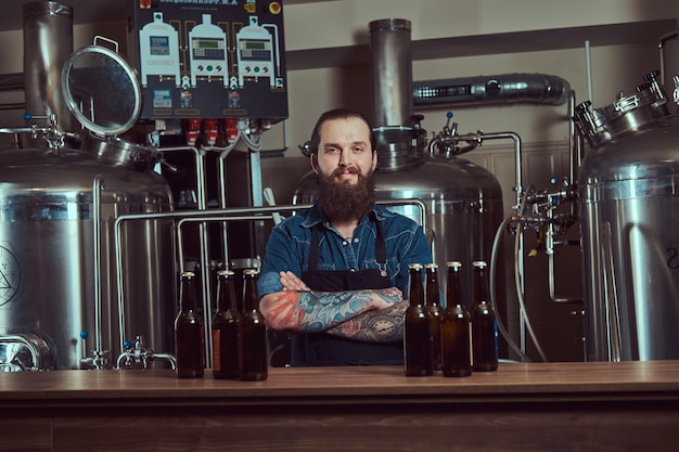 Sonriente hombre hipster tatuado con barba en una camisa de jeans y delantal trabajando en una fábrica de cerveza, de pie con los brazos cruzados detrás del mostrador.
