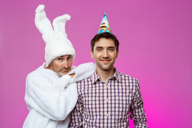 Sonriente hombre y hombre en traje de conejo en la fiesta de cumpleaños sobre pared púrpura.