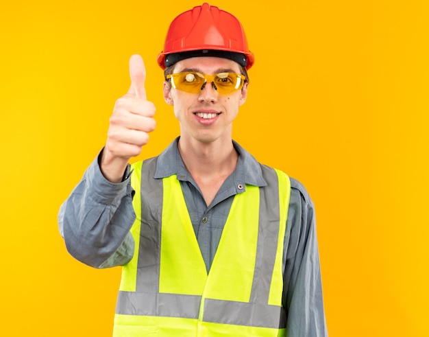 Sonriente a hombre joven constructor en uniforme con gafas mostrando el pulgar hacia arriba