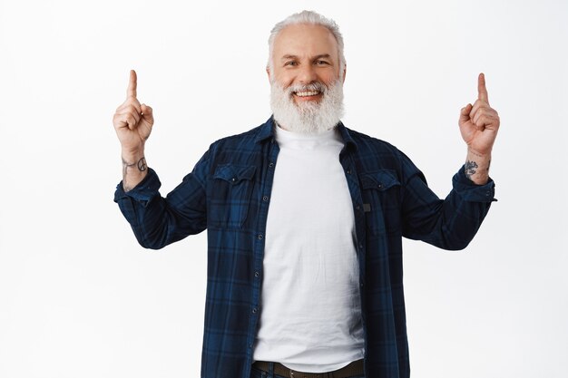 Sonriente hombre mayor feliz apuntando con el dedo hacia arriba y riendo, mostrando y recomendando publicidad, demostrando página web, de pie con ropa elegante contra la pared blanca