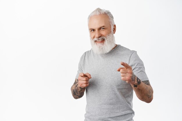 Sonriente hombre mayor guapo con tatuajes que lo invitan, apuntando al frente con expresión de cara feliz, buscando personas, alabando el buen trabajo, de pie sobre una pared blanca