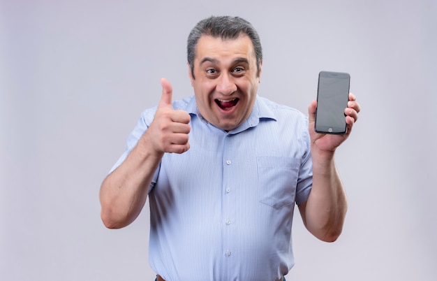 Sonriente hombre de mediana edad con camisa azul sosteniendo teléfono móvil y mostrando los pulgares para arriba mientras está de pie sobre un fondo blanco.