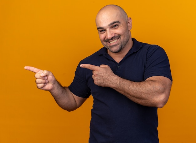 Sonriente hombre de mediana edad casual apuntando al lado aislado en la pared naranja