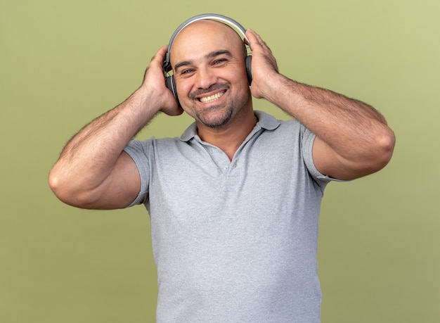 Sonriente hombre de mediana edad casual con audífonos manteniendo las manos sobre ellos mirando al frente aislado en la pared verde oliva