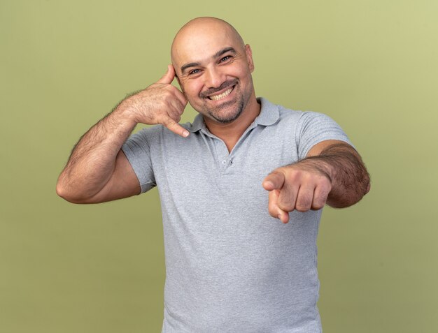 Sonriente hombre de mediana edad casual haciendo gesto de llamada mirando y apuntando al frente aislado en la pared verde oliva