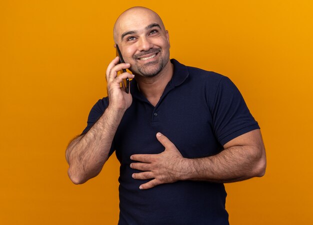 Sonriente hombre de mediana edad casual manteniendo la mano sobre el vientre mirando hacia arriba hablando por teléfono aislado en la pared naranja