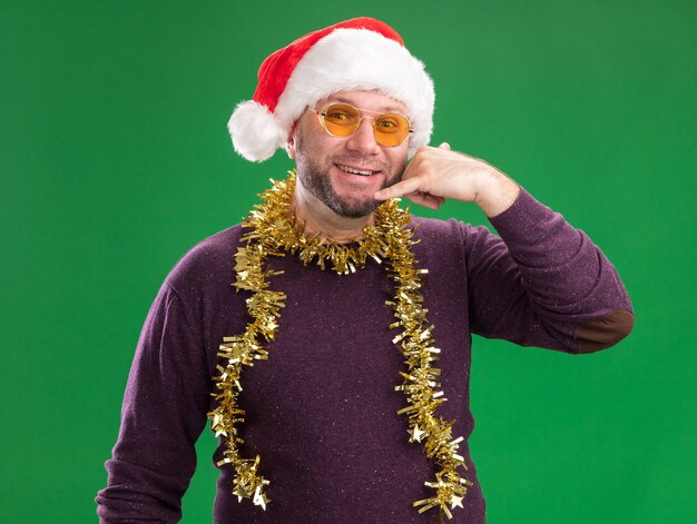 Sonriente hombre de mediana edad con gorro de Papá Noel y guirnalda de oropel alrededor del cuello con gafas mirando a la cámara haciendo gesto de llamada aislado sobre fondo verde