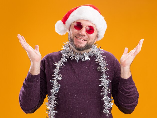 Sonriente hombre de mediana edad con gorro de Papá Noel y guirnalda de oropel alrededor del cuello con gafas mostrando las manos vacías aisladas en la pared naranja