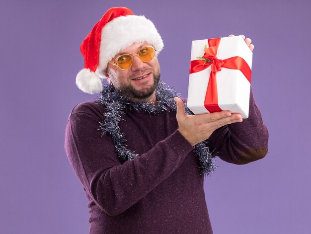 Sonriente hombre de mediana edad con gorro de Papá Noel y guirnalda de oropel alrededor del cuello con gafas con paquete de regalo aislado en la pared púrpura