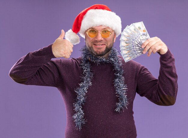 Sonriente hombre de mediana edad con gorro de Papá Noel y guirnalda de oropel alrededor del cuello con gafas sosteniendo dinero mostrando el pulgar hacia arriba aislado en la pared púrpura