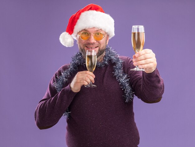 Sonriente hombre de mediana edad con gorro de Papá Noel y guirnalda de oropel alrededor del cuello con gafas sosteniendo dos copas de champán estirando uno aislado en la pared púrpura