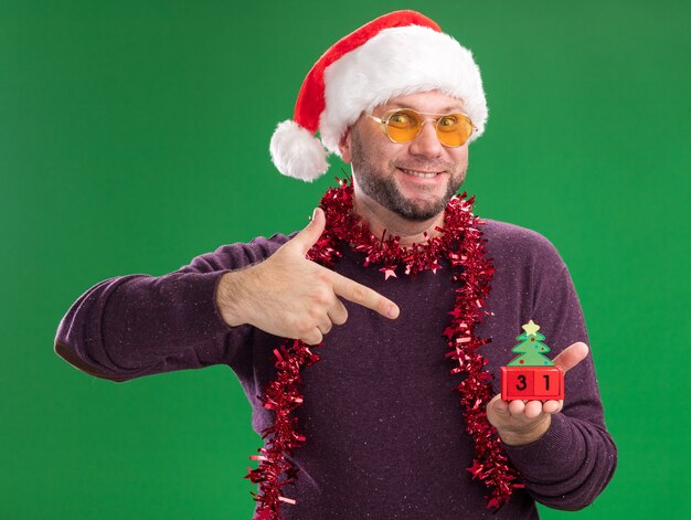 Sonriente hombre de mediana edad con gorro de Papá Noel y guirnalda de oropel alrededor del cuello con gafas sosteniendo y mirando el juguete del árbol de Navidad con fecha mirando a cámara aislada sobre fondo verde