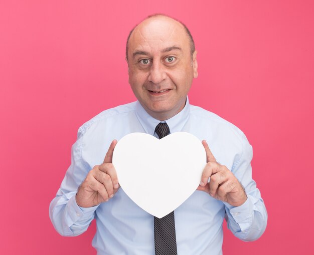 Sonriente hombre de mediana edad vestido con camiseta blanca con corbata sosteniendo una caja en forma de corazón aislada en la pared rosa