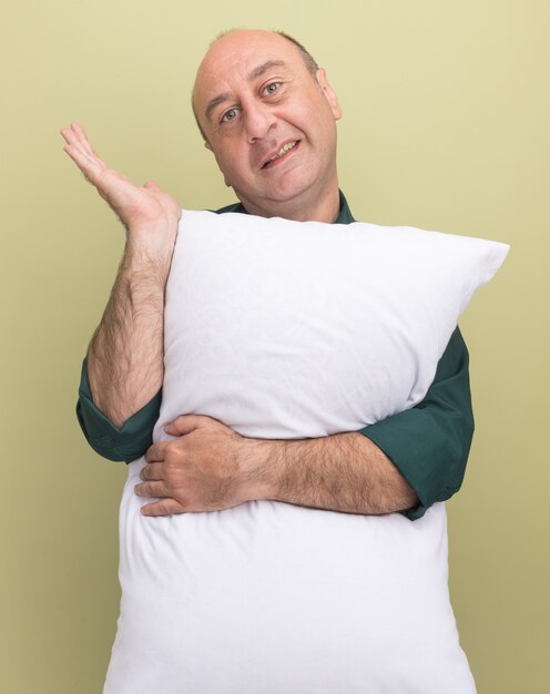 Sonriente hombre de mediana edad vestido con camiseta verde abrazó puntos de almohada con la mano al lado aislado en la pared verde oliva