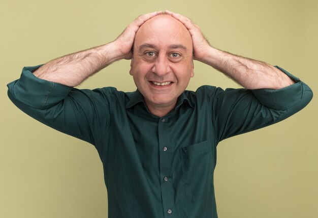 Sonriente hombre de mediana edad vestido con camiseta verde agarró la cabeza aislada en la pared verde oliva