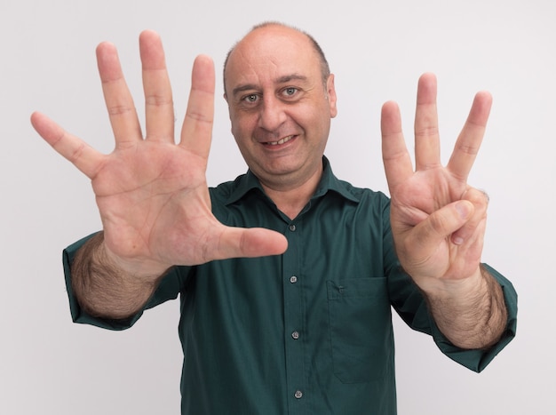 Sonriente hombre de mediana edad vestido con camiseta verde mostrando diferentes números aislados en la pared blanca