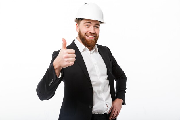 Sonriente hombre de negocios con barba en casco protector mostrando el pulgar hacia arriba