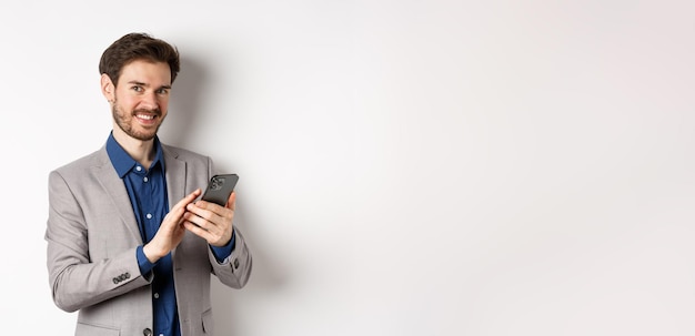 Foto gratuita sonriente hombre de negocios exitoso usando teléfono inteligente sobre fondo blanco de pie en traje