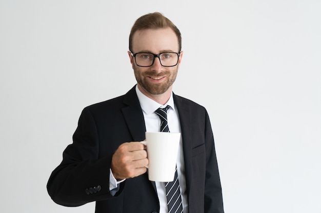 Sonriente hombre de negocios guapo sosteniendo la taza, beber té y mirando a cámara.