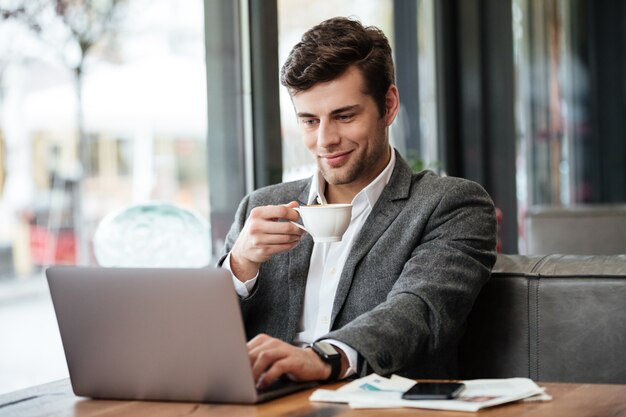 Sonriente hombre de negocios sentado junto a la mesa en la cafetería y usando la computadora portátil