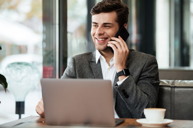 Sonriente hombre de negocios sentado en la mesa de café con computadora portátil mientras habla por teléfono inteligente