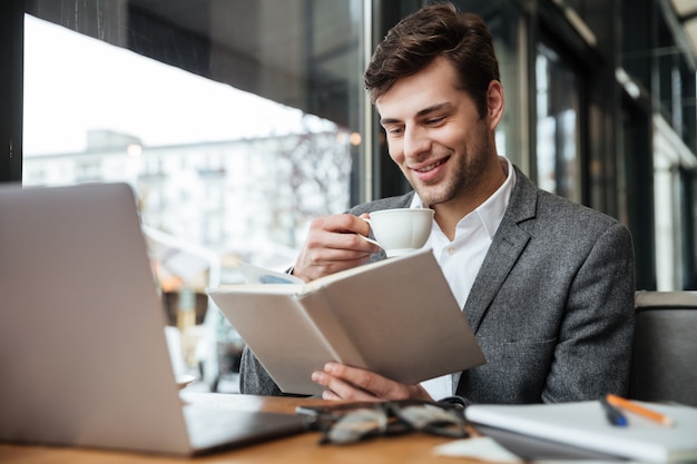 Sonriente hombre de negocios sentado en la mesa de café con computadora portátil mientras lee el libro y bebe café