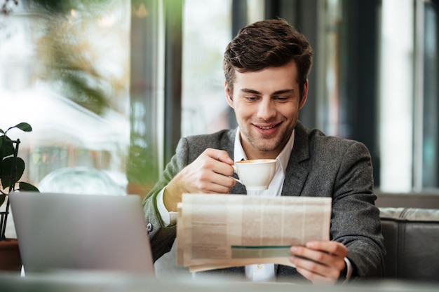 Sonriente hombre de negocios sentado en la mesa de café con computadora portátil mientras lee el periódico y bebe café