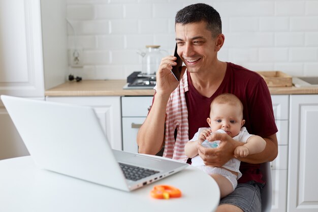 Sonriente hombre positivo vestido con camiseta casual granate sentado en la cocina frente a la computadora portátil y hablando por teléfono, dando agua de la botella para su hija, trabajando en línea.