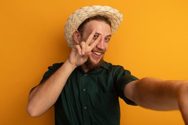 Sonriente hombre rubio guapo con sombrero de playa hace gestos con el signo de la mano de la victoria y pretende sostener el frente aislado en la pared naranja