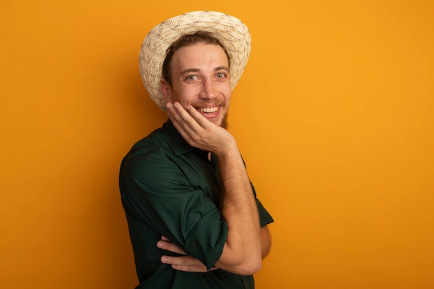 Sonriente hombre rubio guapo con sombrero de playa pone la mano en la cara aislada en la pared naranja