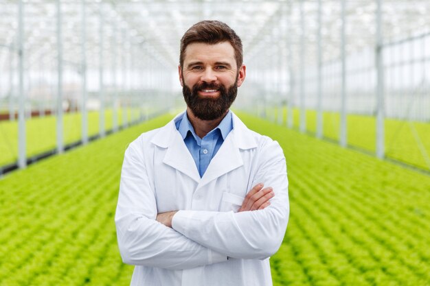 Sonriente investigador masculino con barba se encuentra ante las plantas en un invernadero