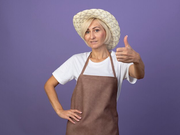 Sonriente jardinero rubio de mediana edad mujer en uniforme con sombrero manteniendo la mano en la cintura mostrando el pulgar hacia arriba aislado en la pared púrpura