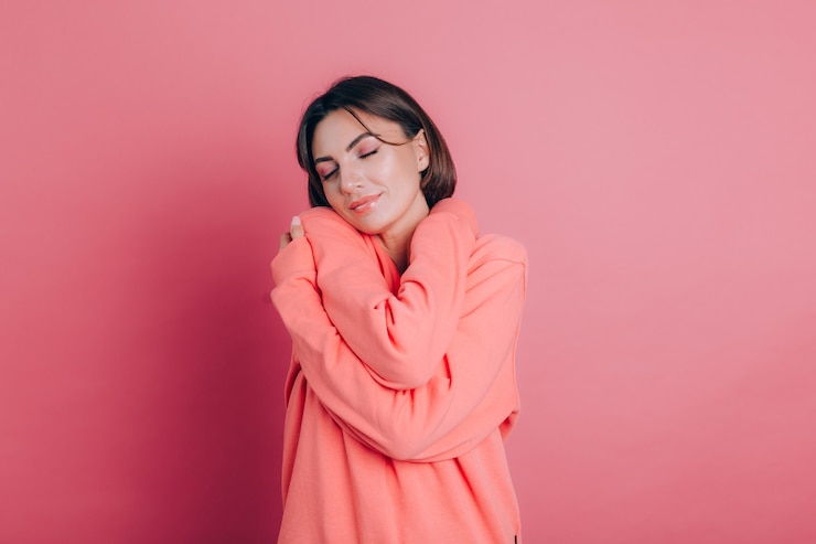 Foto gratuito sonriente joven 20 años en ropa casual aislado en retrato de estudio de fondo rosa. concepto de amor. manteniendo los ojos cerrados tomados de las manos cruzadas abrazándose a sí misma