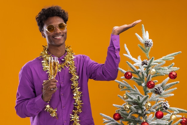Sonriente joven afroamericano con gafas con guirnalda de oropel alrededor del cuello de pie cerca del árbol de Navidad decorado sosteniendo una copa de champán