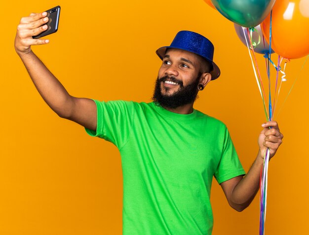 Sonriente joven afroamericano con gorro de fiesta sosteniendo globos tomar un selfie