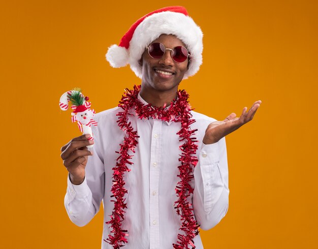 Sonriente joven afroamericano con gorro de Papá Noel y gafas con guirnalda de oropel alrededor del cuello sosteniendo adorno de bastón de caramelo mirando a cámara mostrando la mano vacía aislada sobre fondo naranja