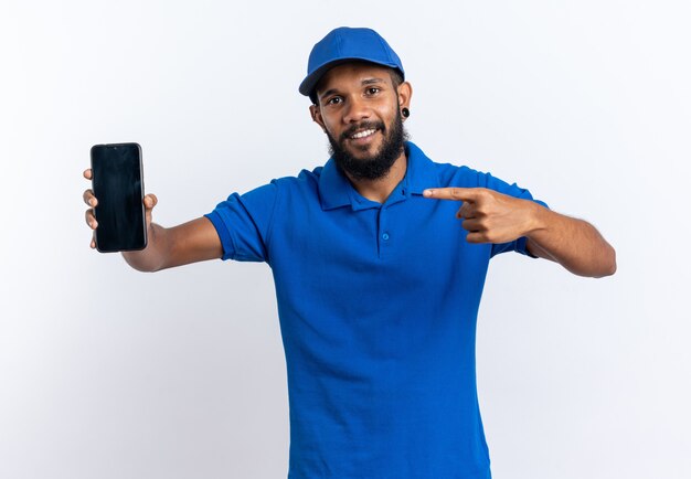 Sonriente joven afroamericano repartidor sosteniendo y apuntando al teléfono aislado sobre fondo blanco con espacio de copia