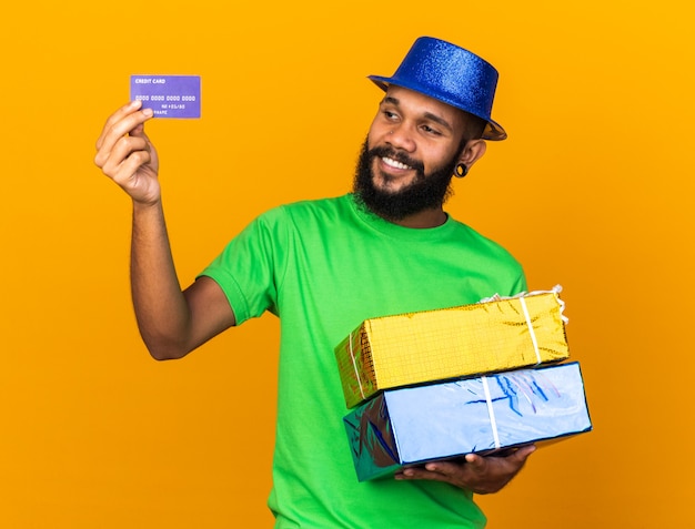 Sonriente joven afroamericano con sombrero de fiesta sosteniendo cajas de regalo y mirando la tarjeta de crédito aislada en la pared naranja