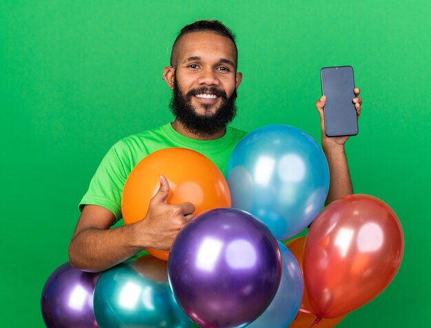 Sonriente joven afroamericano vistiendo camiseta verde de pie detrás de globos sosteniendo teléfono