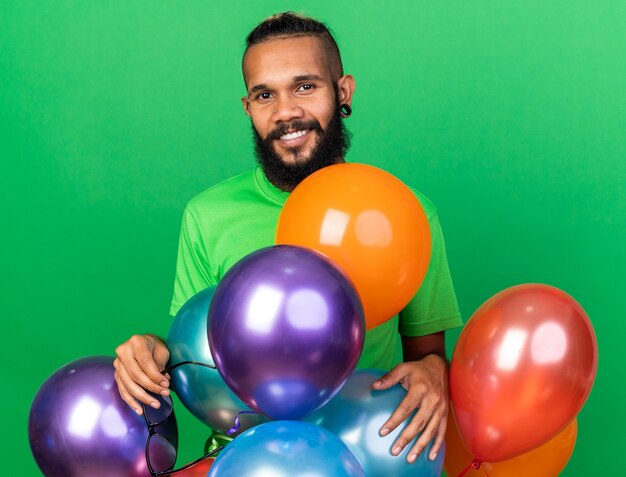 Sonriente joven afroamericano vistiendo camiseta verde de pie detrás de globos