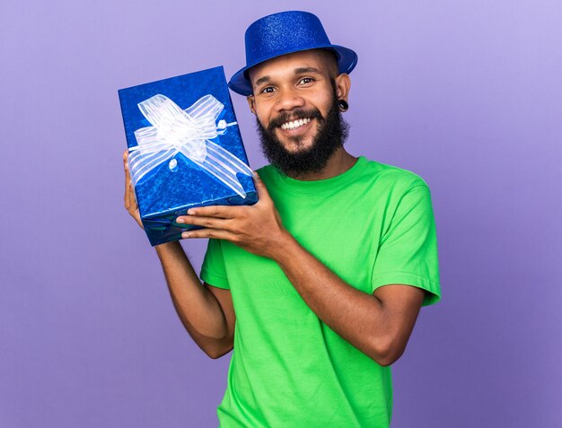 Sonriente joven afroamericano vistiendo gorro de fiesta con caja de regalo aislada en la pared azul