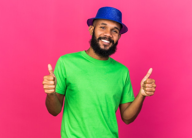 Sonriente joven afroamericano vistiendo gorro de fiesta mostrando los pulgares para arriba aislados en la pared rosa