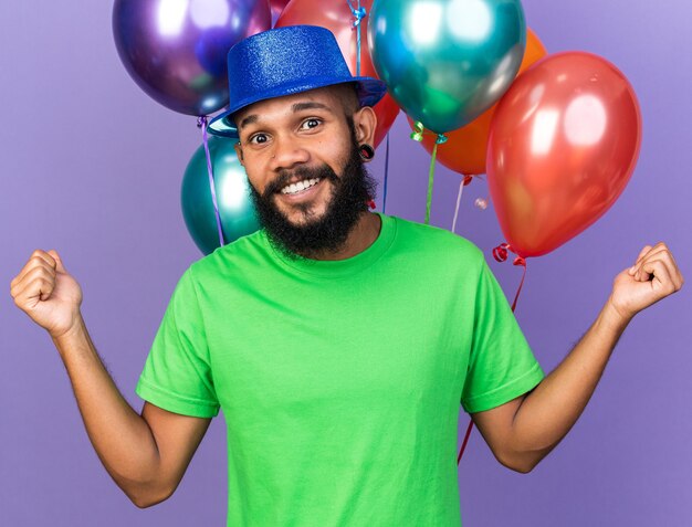 Sonriente joven afroamericano vistiendo gorro de fiesta de pie delante de globos extendiendo las manos