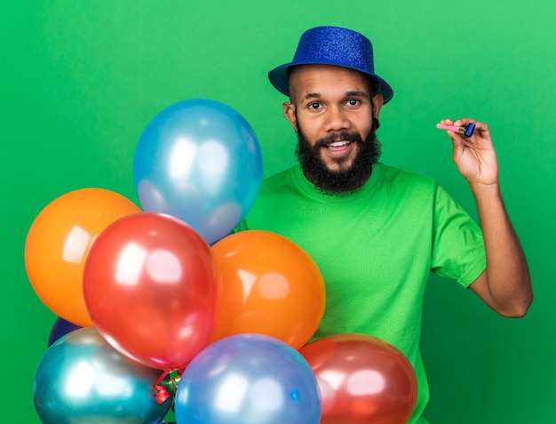 Foto gratuita sonriente joven afroamericano vistiendo gorro de fiesta sosteniendo globos con silbato de fiesta aislado en la pared verde