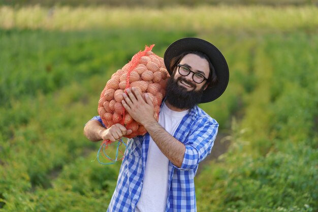 Sonriente joven agricultor sosteniendo un saco de patatas frescas en el campo de patatas verdes