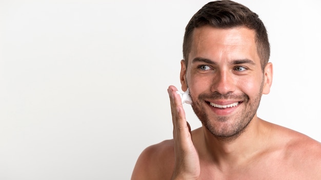 Sonriente joven aplicando espuma de afeitar sobre fondo blanco