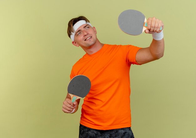 Sonriente joven apuesto deportivo con diadema y muñequeras sosteniendo y levantando raquetas de ping pong y mirando al lado aislado en la pared verde oliva