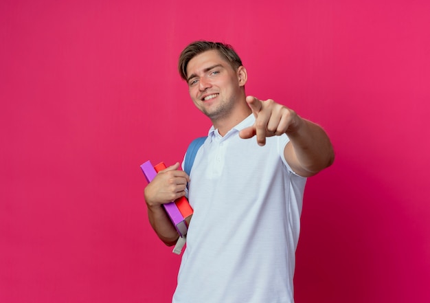 Sonriente joven apuesto estudiante vistiendo bolsa trasera sosteniendo libros