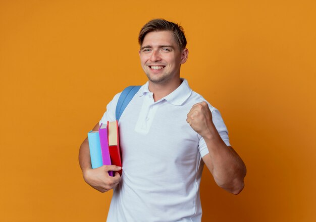 Sonriente joven apuesto estudiante vistiendo bolsa trasera sosteniendo libros