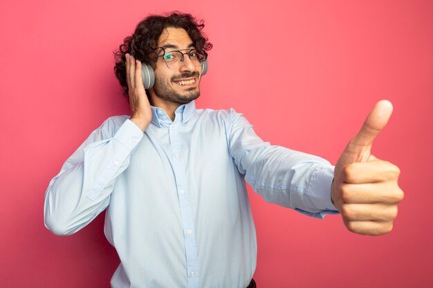 Sonriente joven apuesto hombre caucásico con gafas y auriculares mirando a la cámara poniendo la mano en los auriculares mostrando el pulgar hacia arriba aislado sobre fondo carmesí